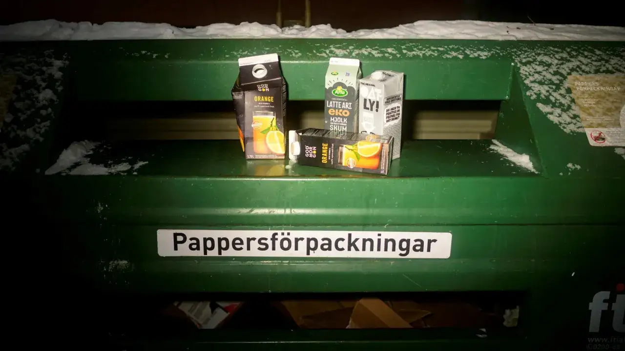 Tre tomma dryckeskartonger, en med apelsinmotiv, ligger redo f&ouml;r kartong &aring;tervinning i en gr&ouml;n beh&aring;llare.