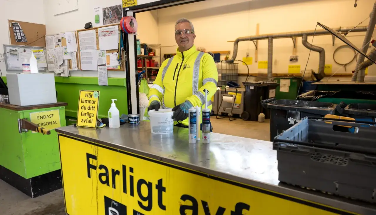 Man i varselkläder hanterar farligt avfall vid en återvinningsstation. Skyltar visar att det är för personal och att farligt avfall lämnas här.