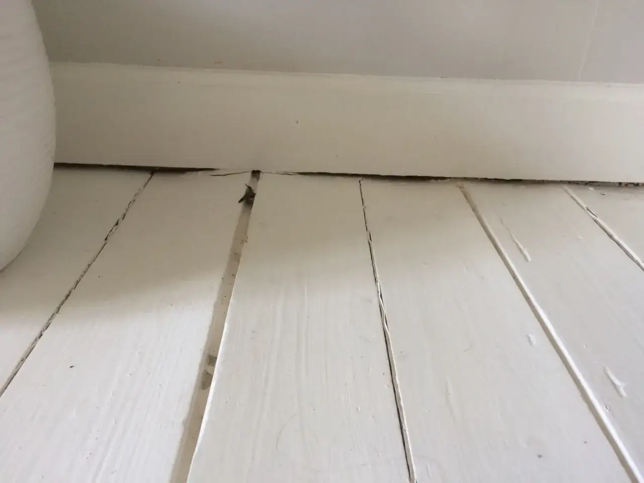 Vita träplankor med sprickor och ojämnheter, redo att slipa målat golv för en jämnare yta.