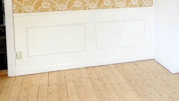 Ett rum med en blommig tapet, vitmålad panel och ett trägolv som väntar på att behandlas med såpa med linolja.